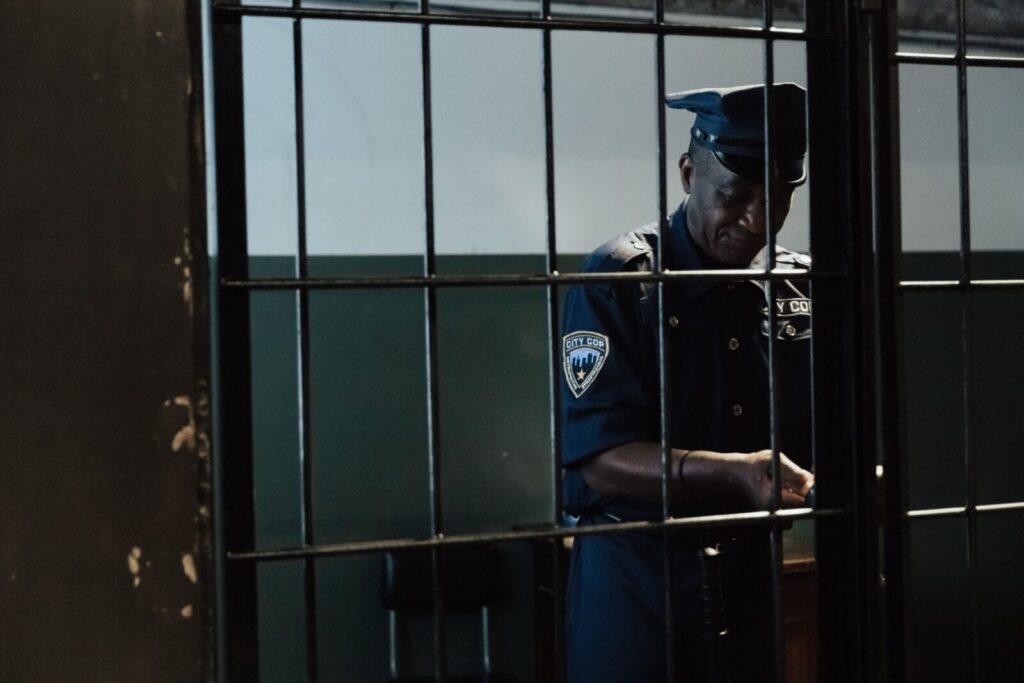 A security officer in uniform standing behind bars in a dimly lit room, evoking themes of law enforcement and confinement.