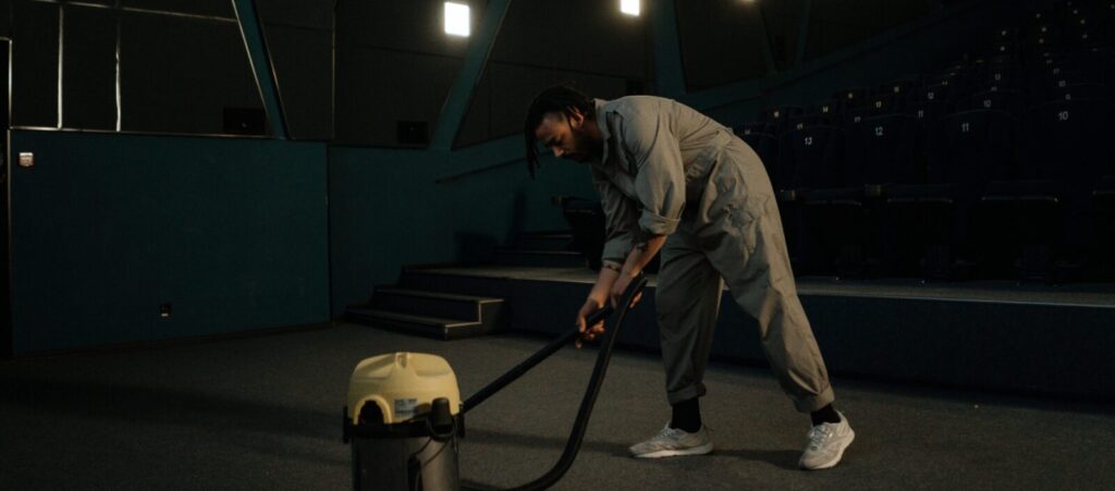 A cinema employee cleaning the theater floor with a vacuum cleaner under dim lights.