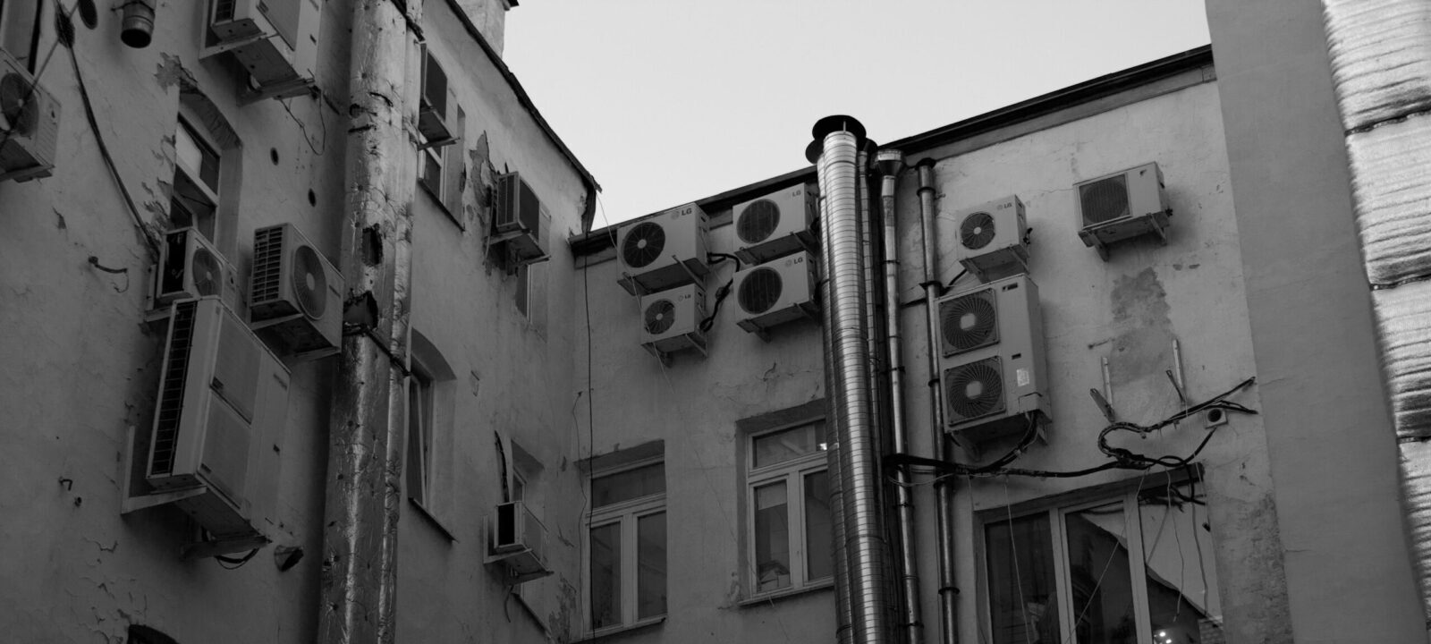 A black and white image of urban building exterior with multiple air conditioning units and pipes.
